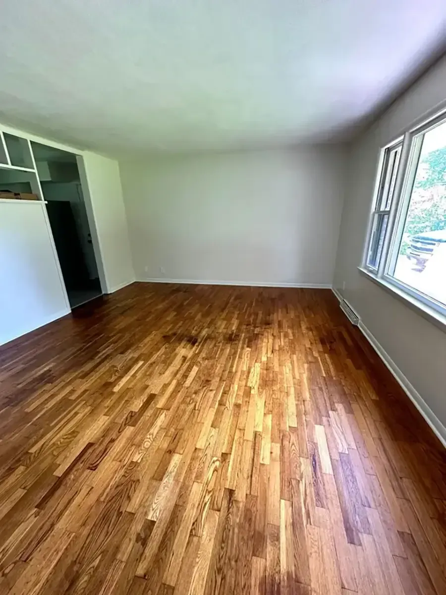 Empty room with hardwood floors, white walls, and a large window letting in sunlight.