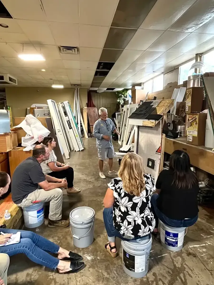 People seated in a cluttered room while a man stands and speaks near shelves and leaning ladders.
