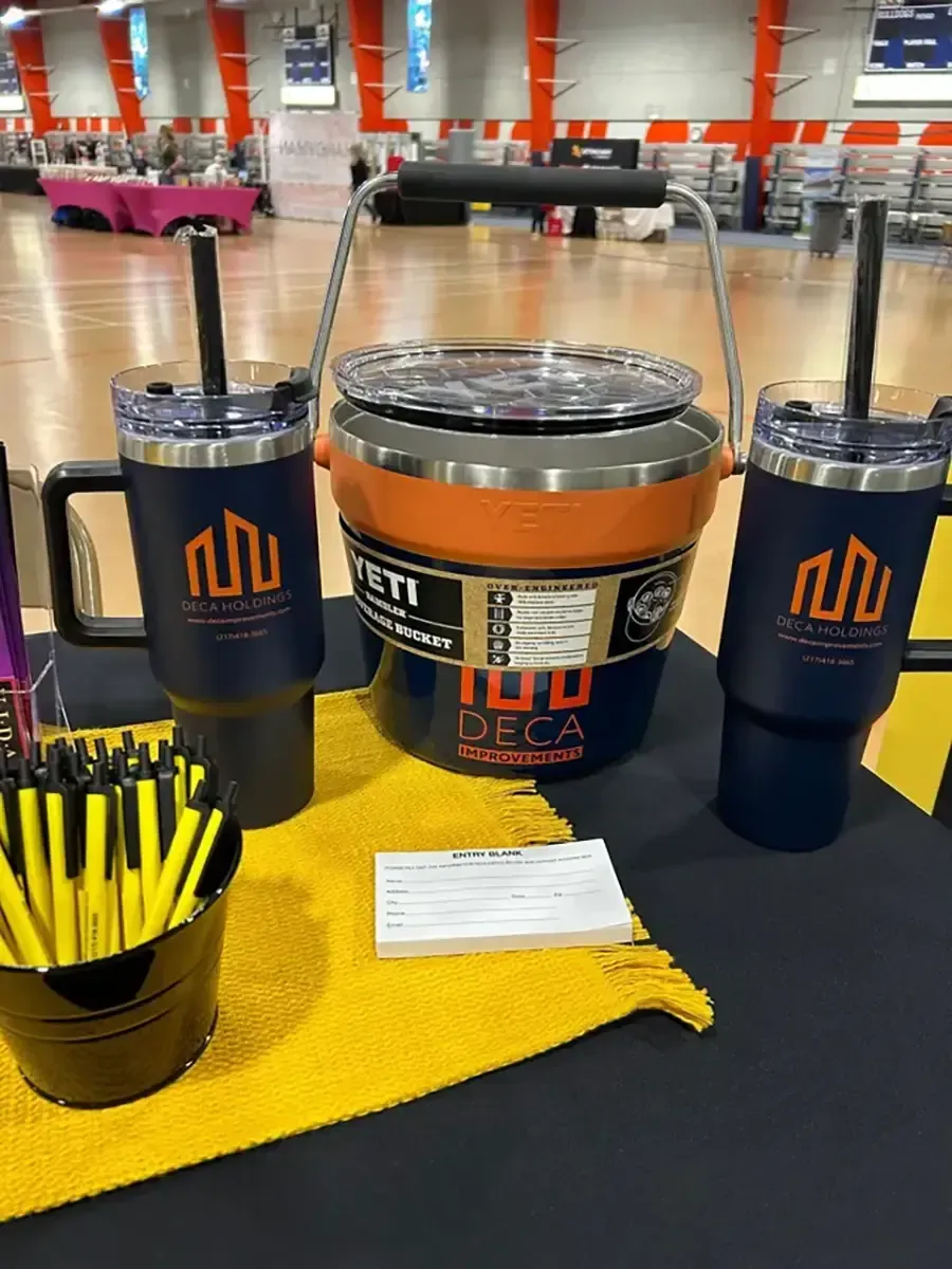 Event display with orange-and-blue drink tumblers, a black bucket of yellow pens, and a cloth on a table
