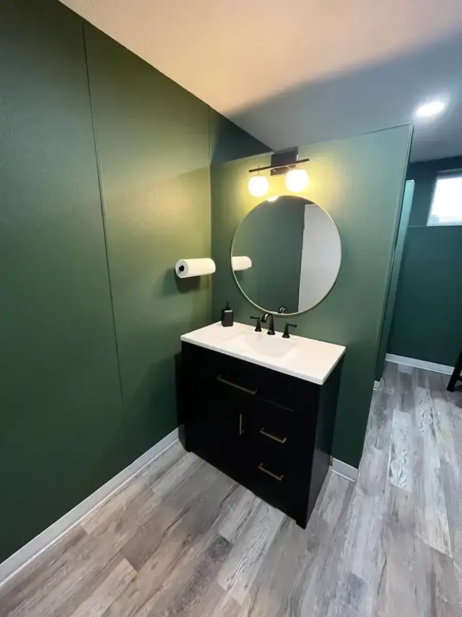 Modern bathroom vanity with round mirror, black cabinet, green walls, and wood-look floor.