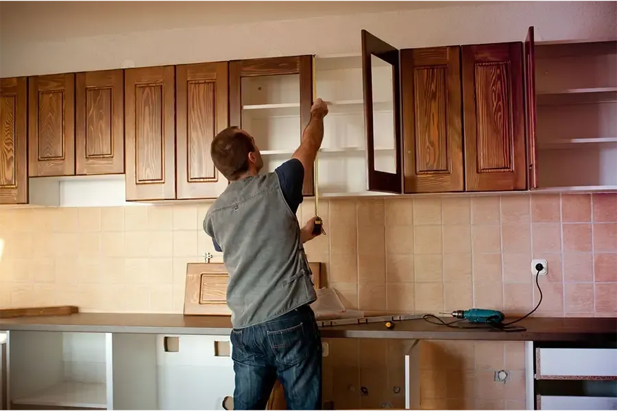Person reaching into an open kitchen cabinet in a room with wood cabinets and beige tile walls.