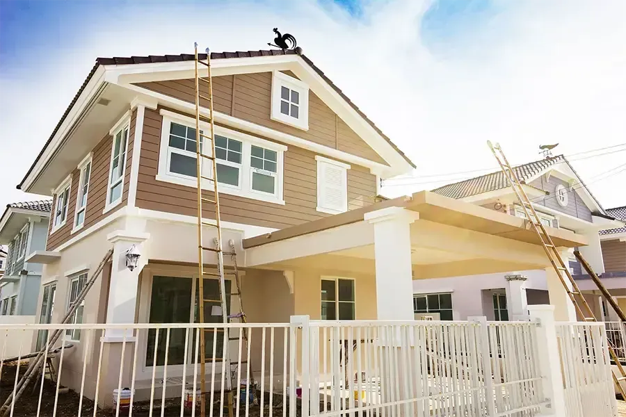 Two-story tan house with white trim and a covered porch, seen behind a white fence and ladders.