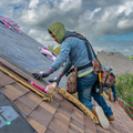 Person kneeling on a shingled roof, installing pink insulation near a skylight under cloudy skies