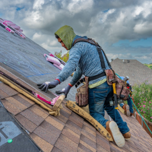 Person kneeling on a shingled roof, installing pink insulation near a skylight under cloudy skies