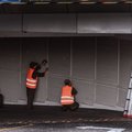 Two workers in orange safety vests inspecting a closed metal shutter in a dim parking garage.
