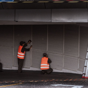 Two workers in orange safety vests inspecting a closed metal shutter in a dim parking garage.