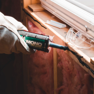 Gloved hand applying black sealant along a window frame beside a windowsill and insulation