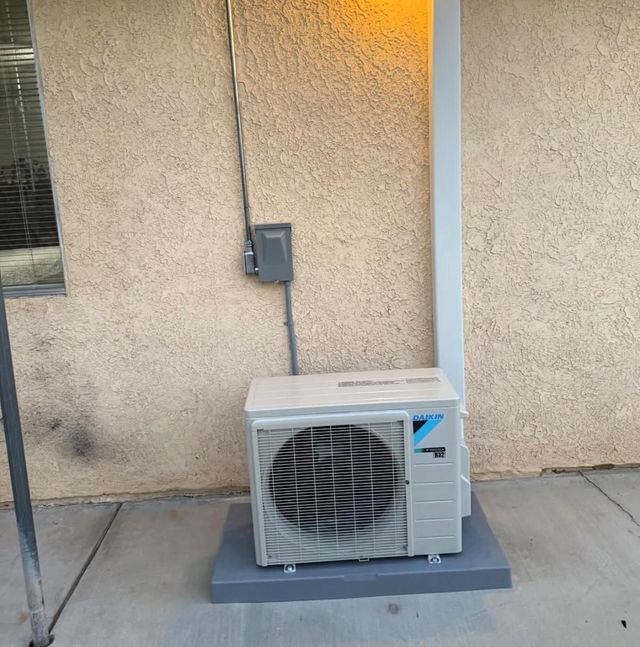 Outdoor air conditioner unit beside a beige stucco wall with a gray electrical box and conduit