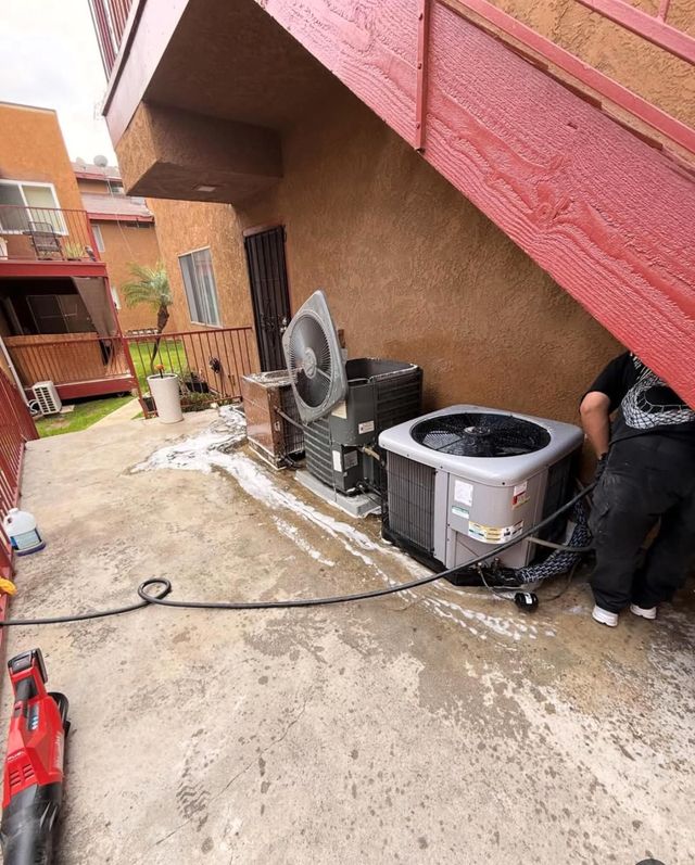 Outdoor apartment walkway with two HVAC units, a fan, and a worker under a stairway on a wet concrete surface