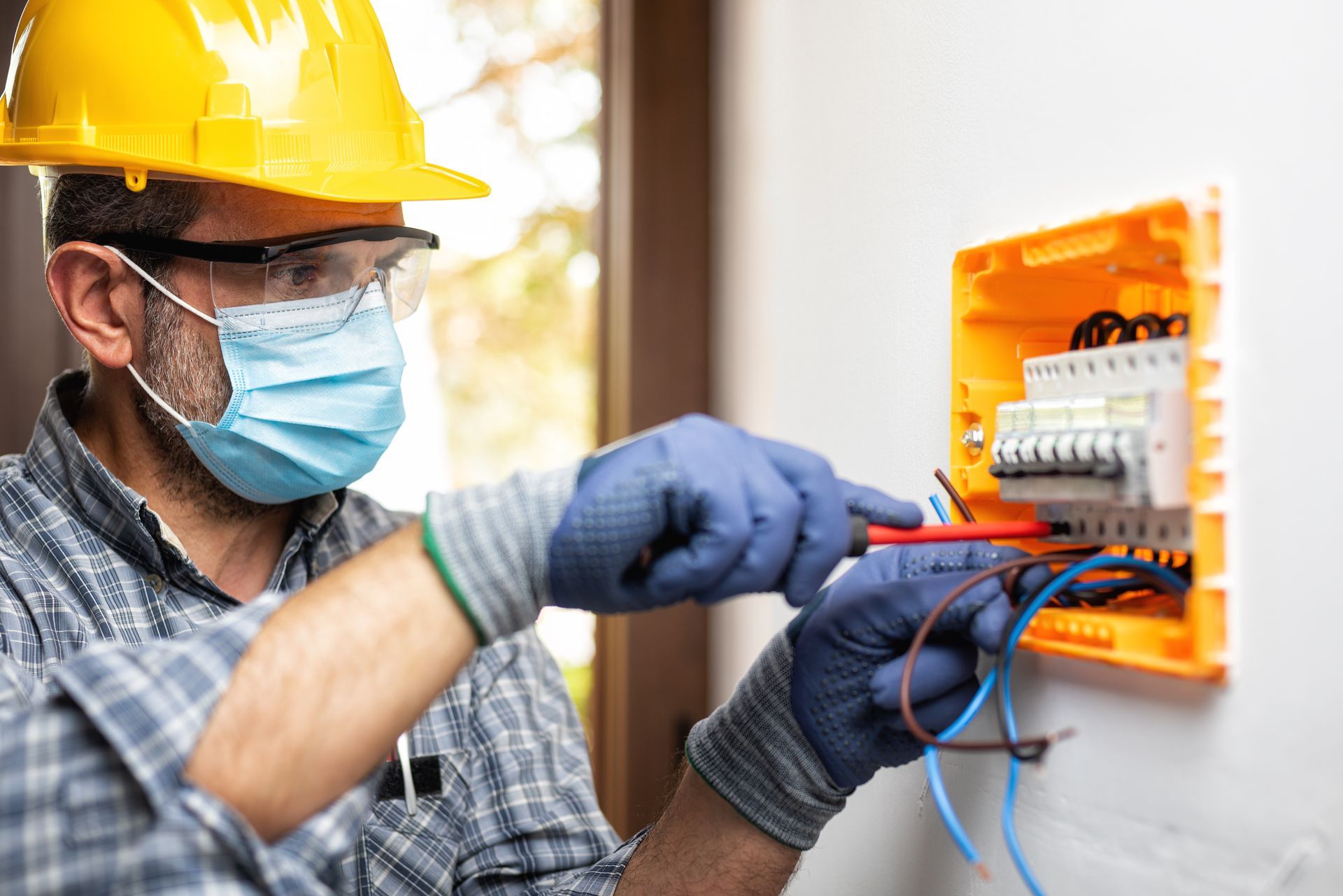 Electrician in hard hat and mask wiring an orange electrical panel indoors