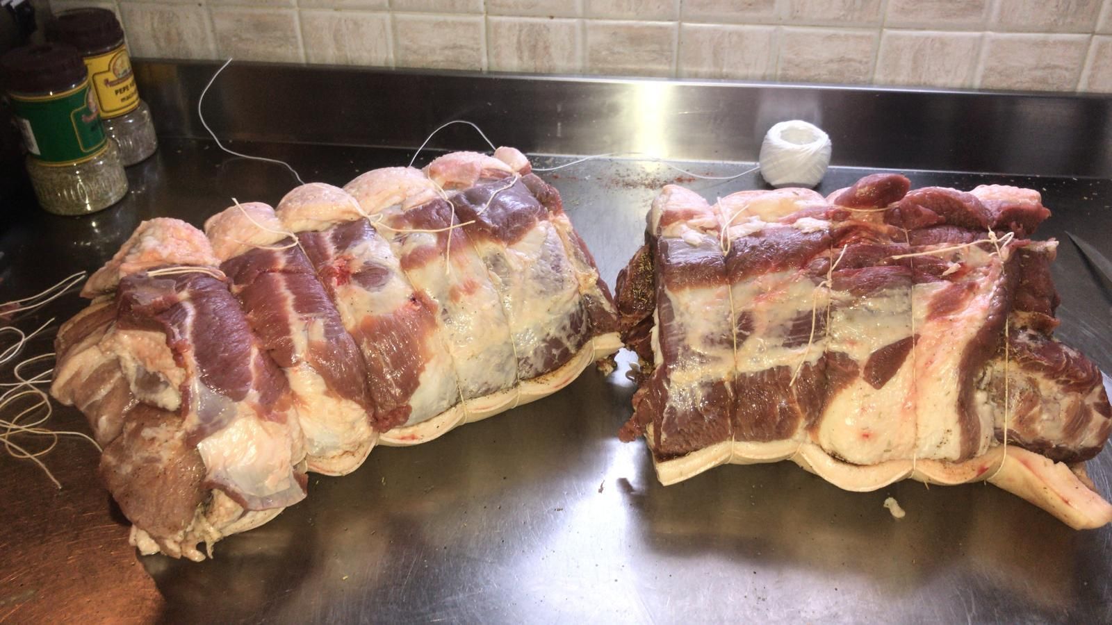Un arrosto di maiale crudo avvolto nella pancetta, tagliato a pezzi, si trova su un piano di lavoro, con un barattolo di spezie nelle vicinanze.