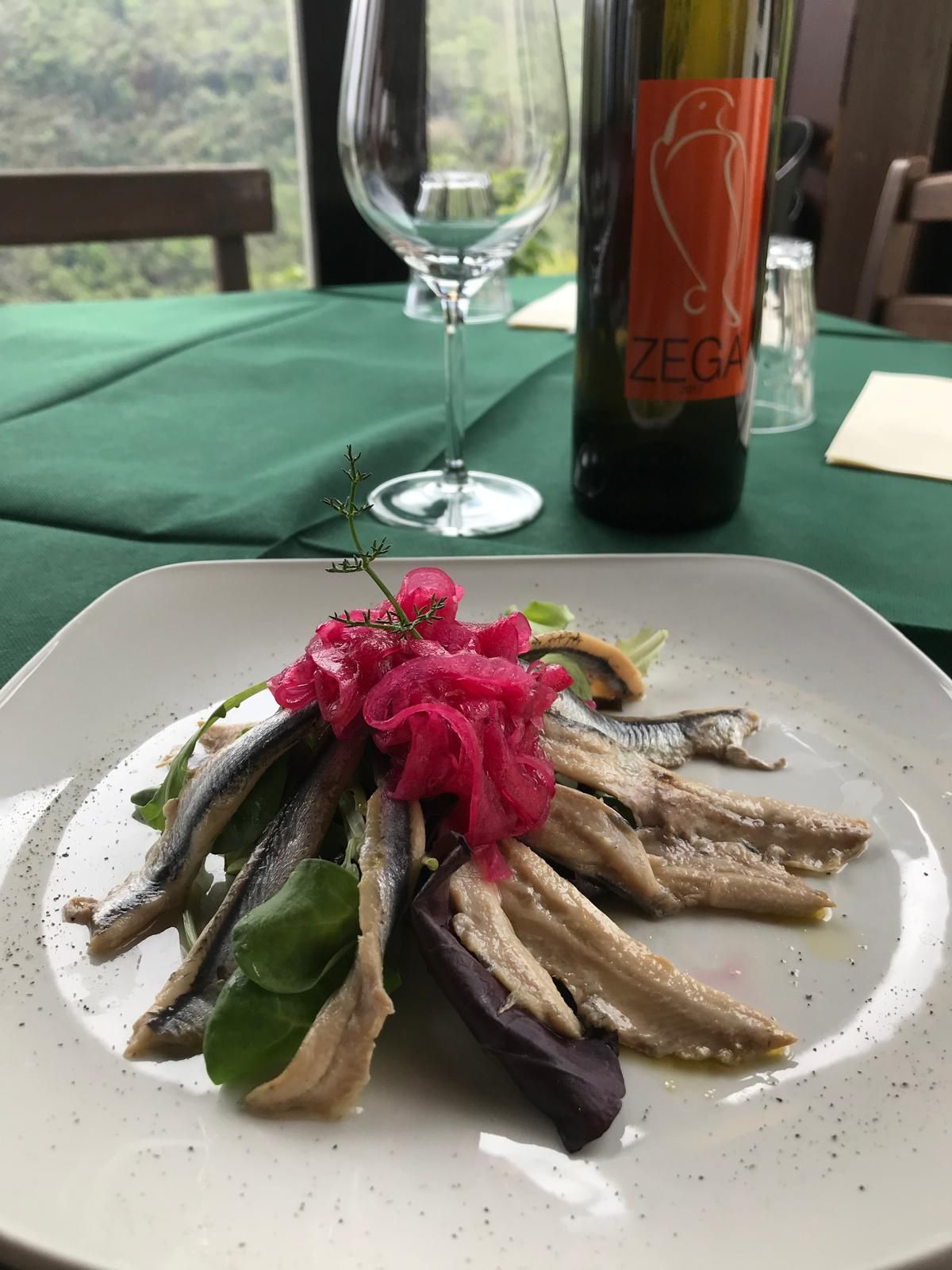 Piatto di pesce con guarnizione rosa su una tovaglia verde, accanto a un bicchiere e una bottiglia di vino.