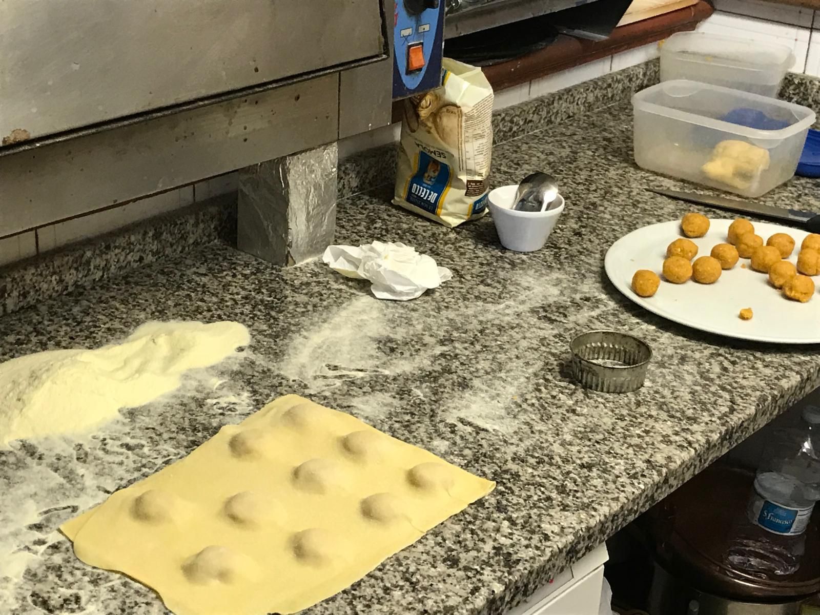 Piano di lavoro infarinato con pasta stesa, quadrati di pasta tagliati e un vassoio di palline di pasta in una cucina