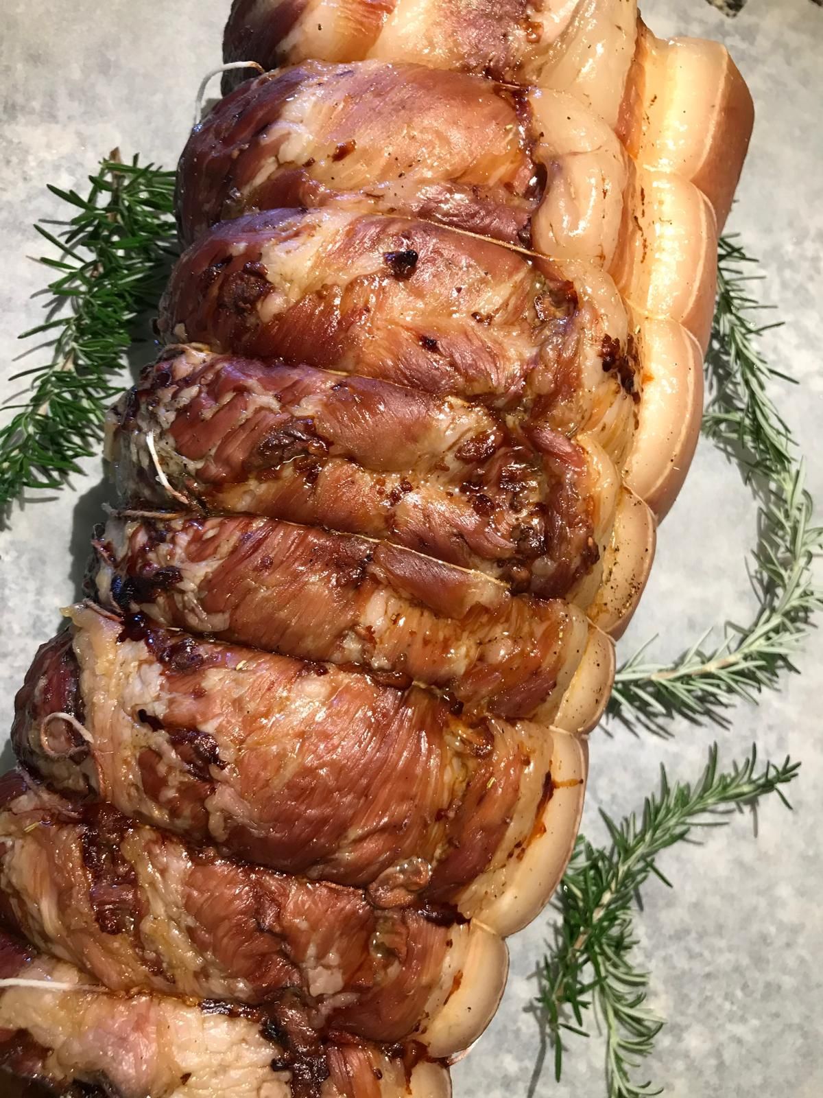 Arrosto di maiale ripieno alla griglia, avvolto nella pancetta e guarnito con rosmarino, servito su un piatto bianco.