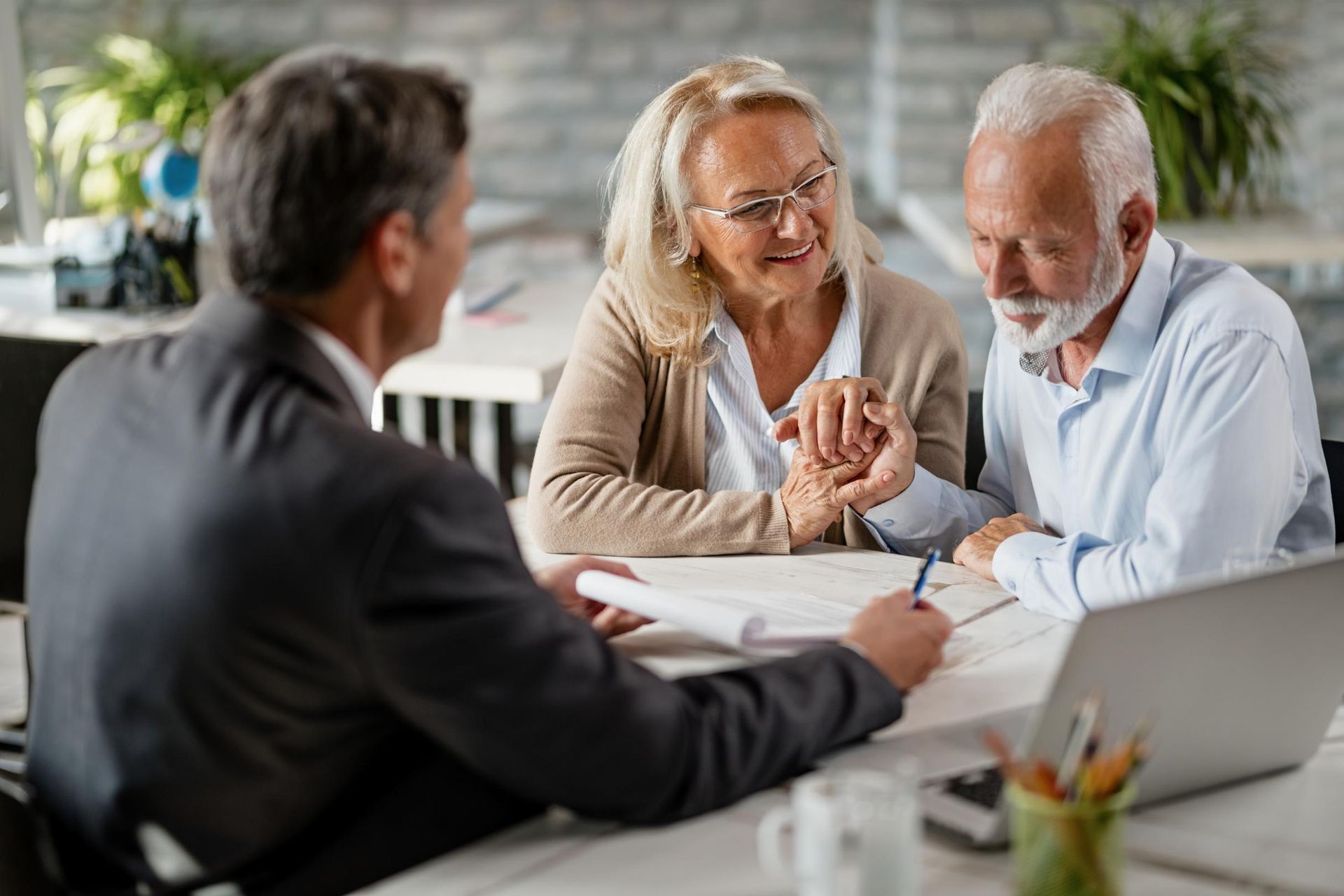 Financial advisor assisting an elderly couple with paperwork at a table; they are holding hands.