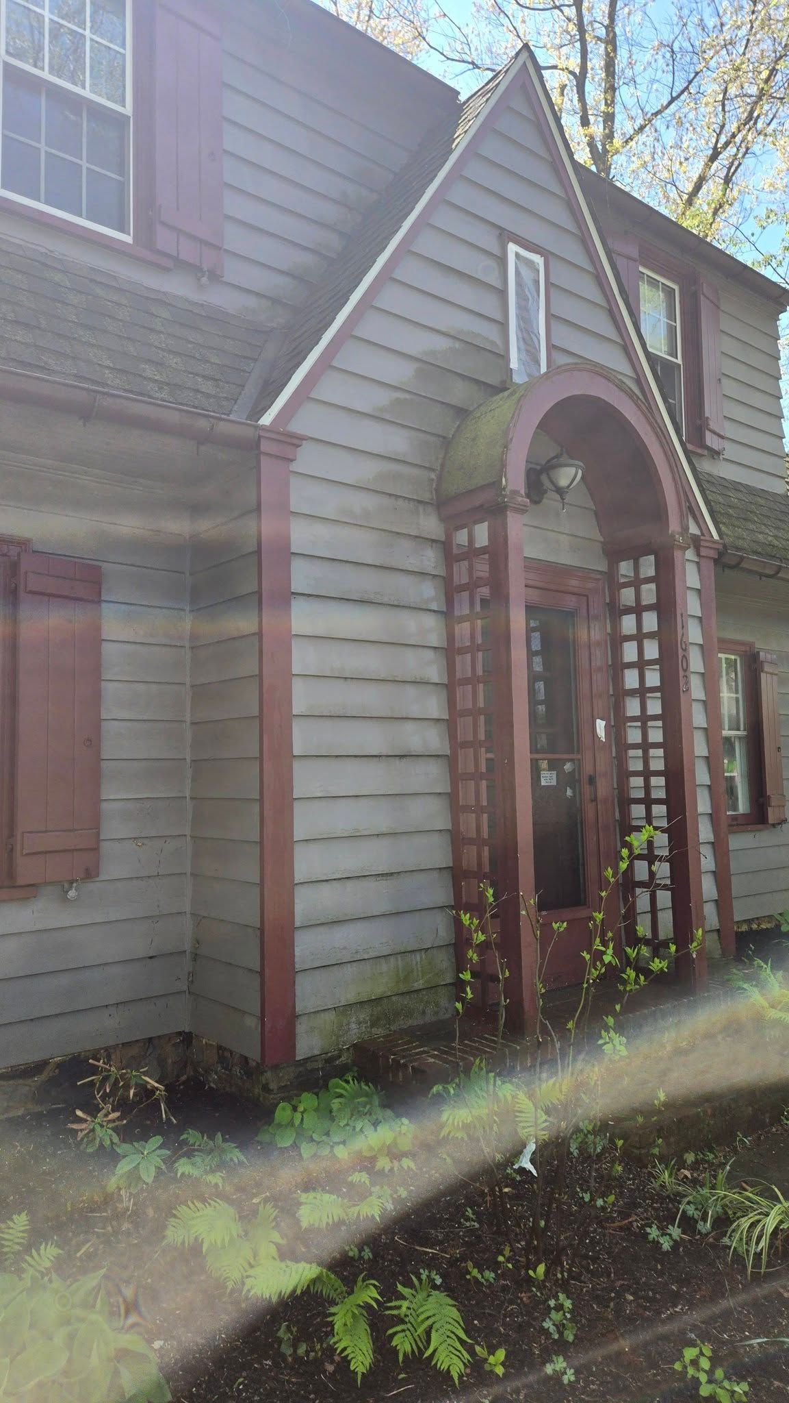 Gray house exterior with a brick arched doorway and sunlit siding, viewed from the front yard