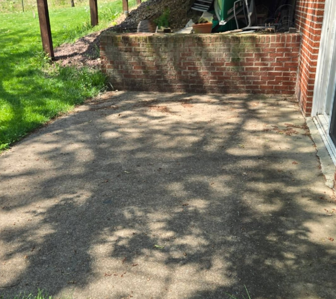 Shaded concrete patio beside a brick wall and steps, with grass and trees in the background