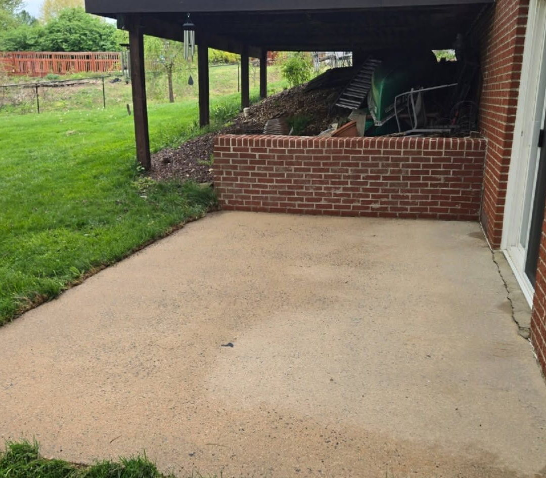 Covered concrete patio beside a brick wall and grassy yard with a storage area under a roofed structure
