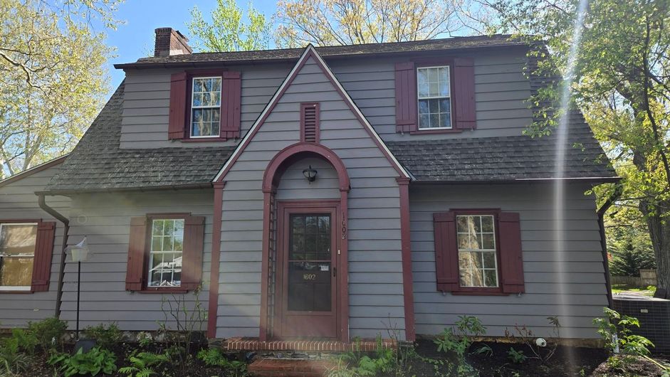 Gray house with red trim, dormer windows, and an arched front door in a leafy yard