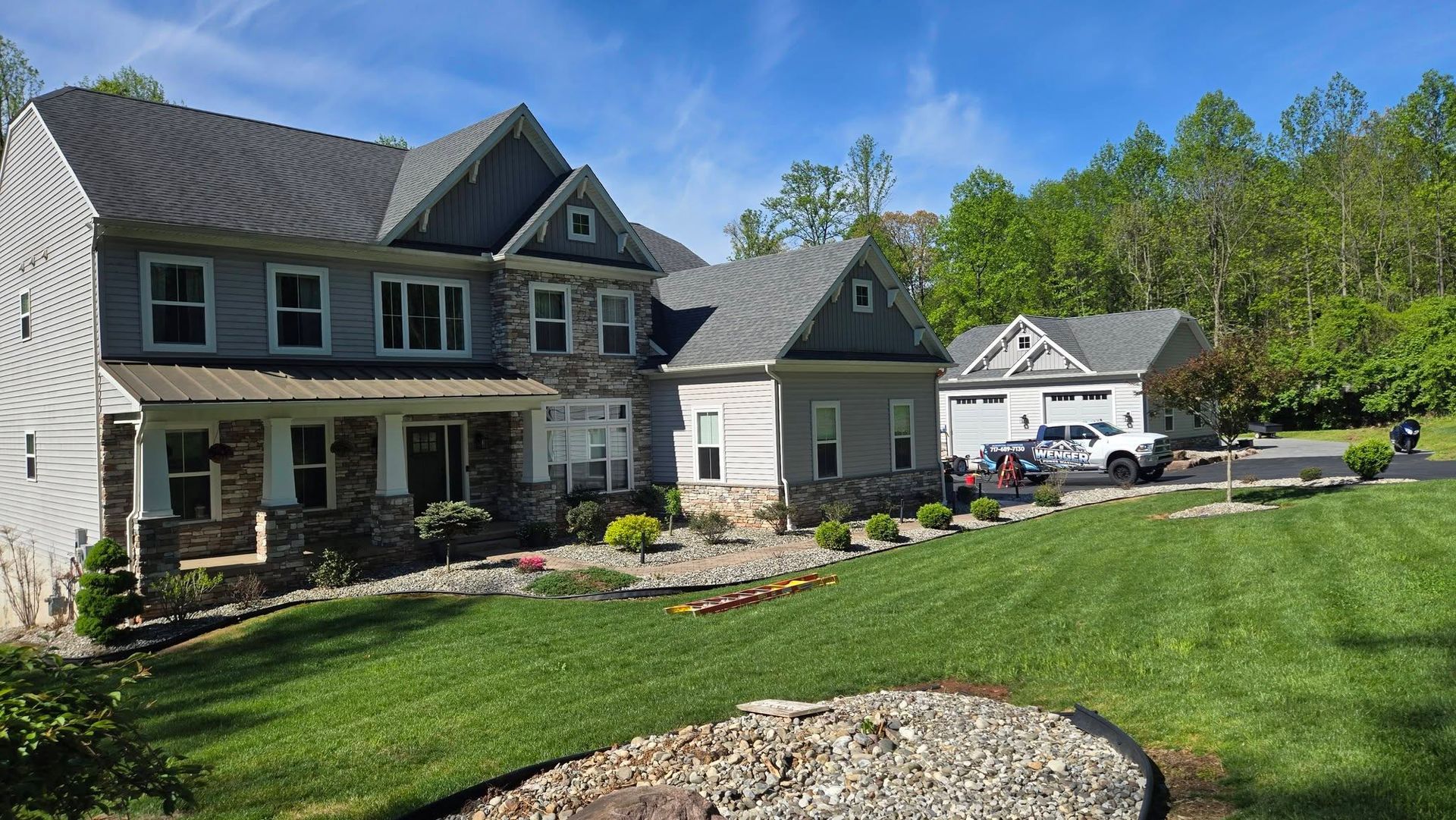 Two-story suburban house with a green lawn, stone landscaping, and parked cars under a blue sky.
