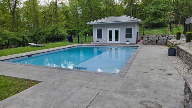 Backyard swimming pool with a concrete deck and gray pool house surrounded by trees