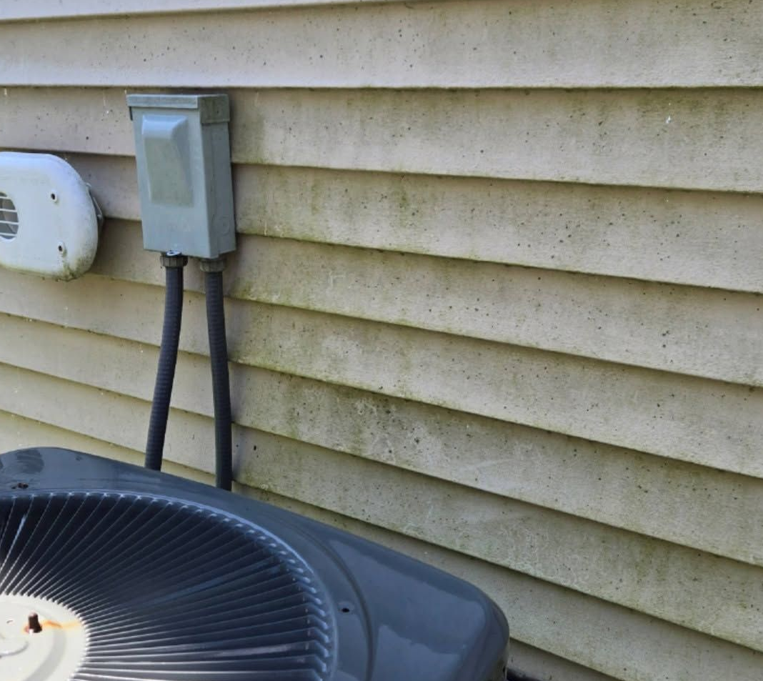 Outdoor air conditioner unit beside a beige house wall with an electrical disconnect box above it