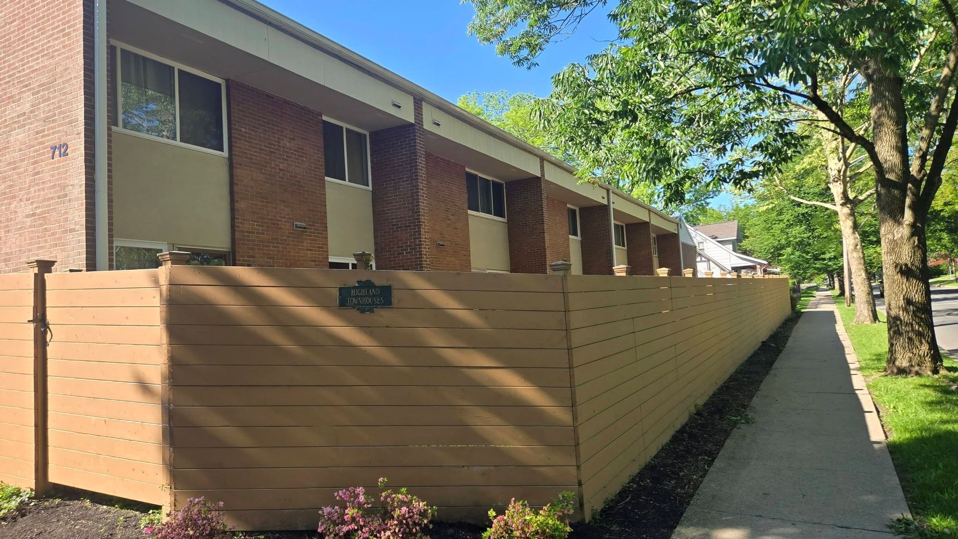 Brick apartment building behind a tan fence along a shaded sidewalk