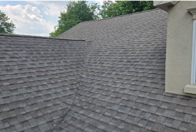 Gray shingled roof with a dormer and a partial view of a beige house against a cloudy sky