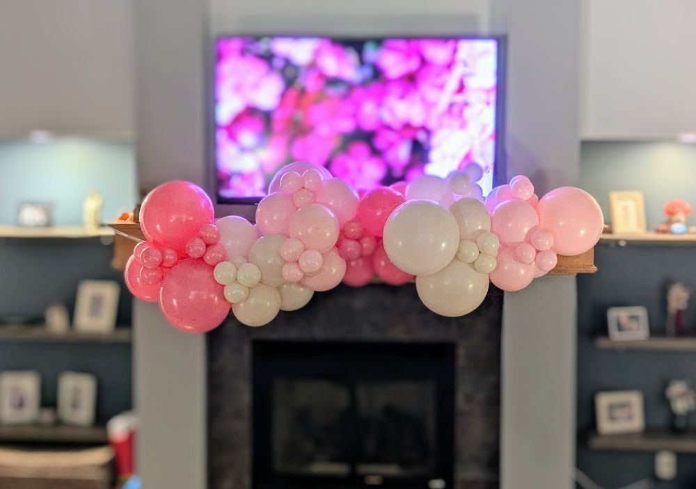 A fireplace mantel decorated with a pink and white balloon garland, with a TV showing pink flowers above.