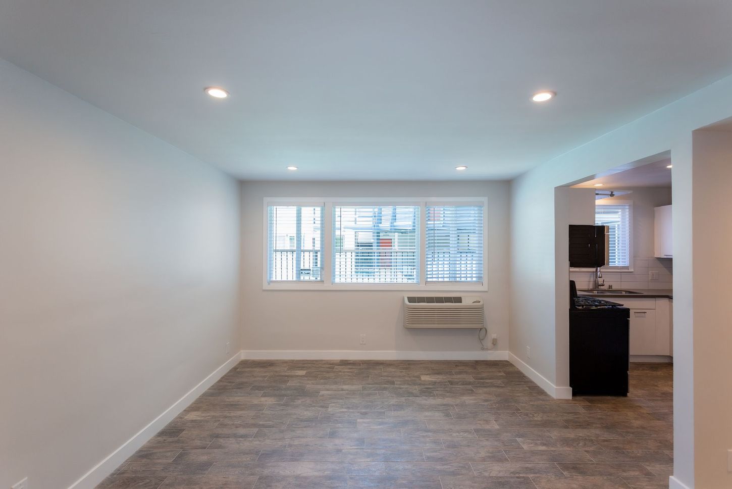 Empty white living room with large window, recessed lights, and a kitchen doorway on the right