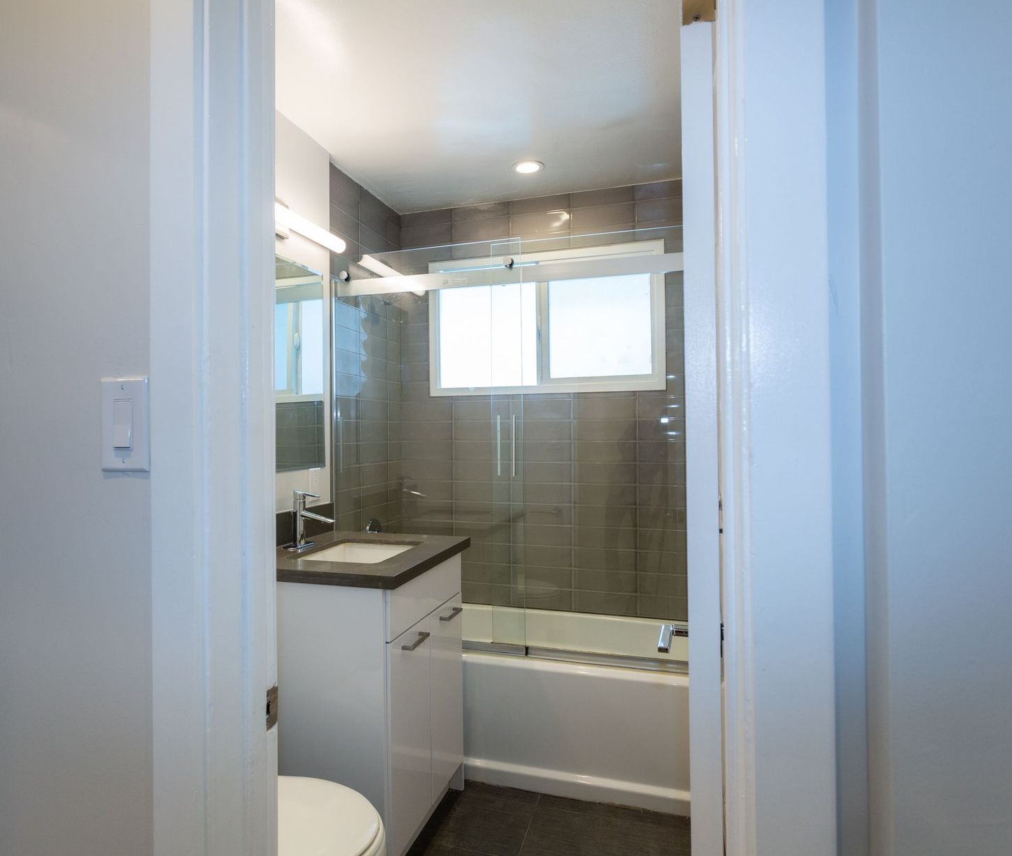 Bathroom vanity and tub viewed through a doorway, with gray tile and a window above the tub.
