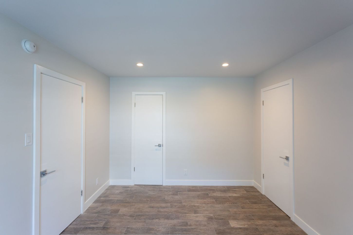 Empty white room with three doors, recessed ceiling lights, and wood-look flooring.