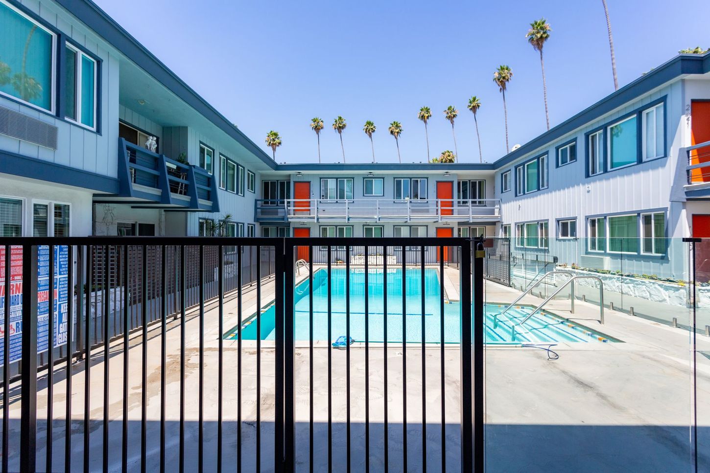 Apartment courtyard with a swimming pool, black fence, and blue sky with palm trees
