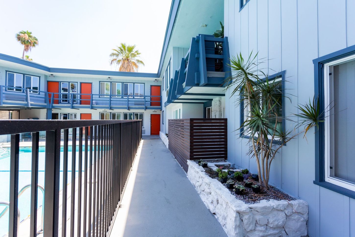 Outdoor apartment walkway with black railings, blue-gray walls, and potted plants in white stone planters.