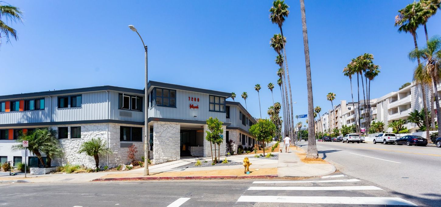 Sunny street corner with modern apartments, palm trees, and parked cars along a wide road