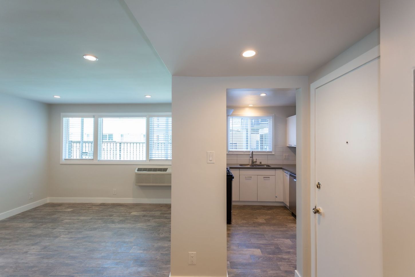 Empty apartment interior with carpeted living area, kitchen nook, white walls, and large windows