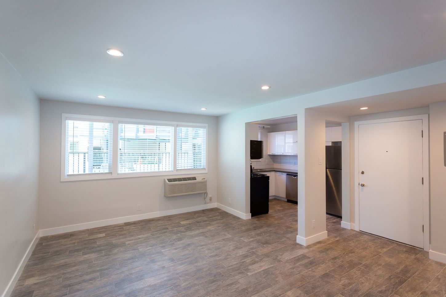 Spacious empty living room with gray flooring, white walls, recessed lights, and kitchen entry.
