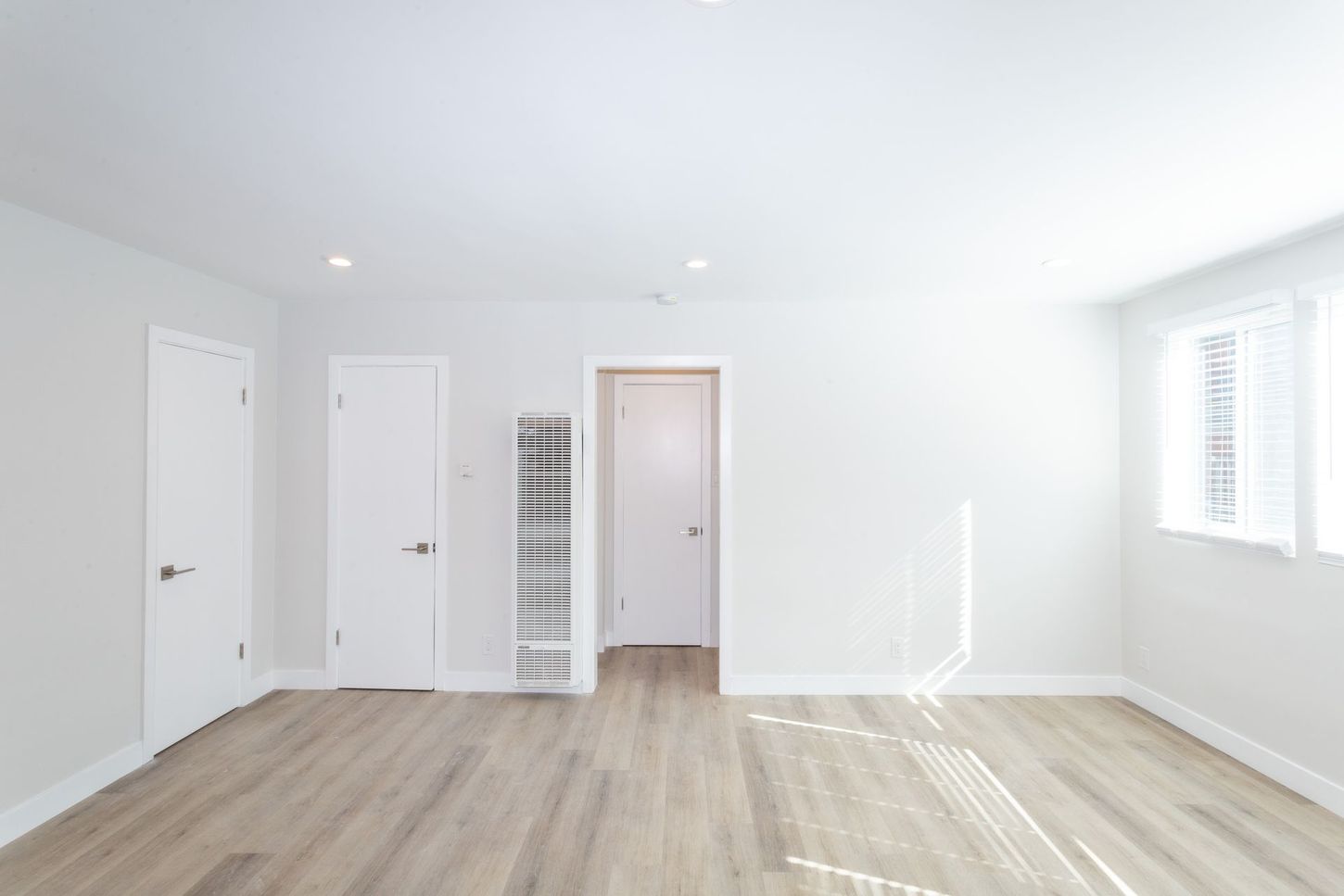 Bright empty bedroom with white walls, light wood floors, doors, and a window with blinds.
