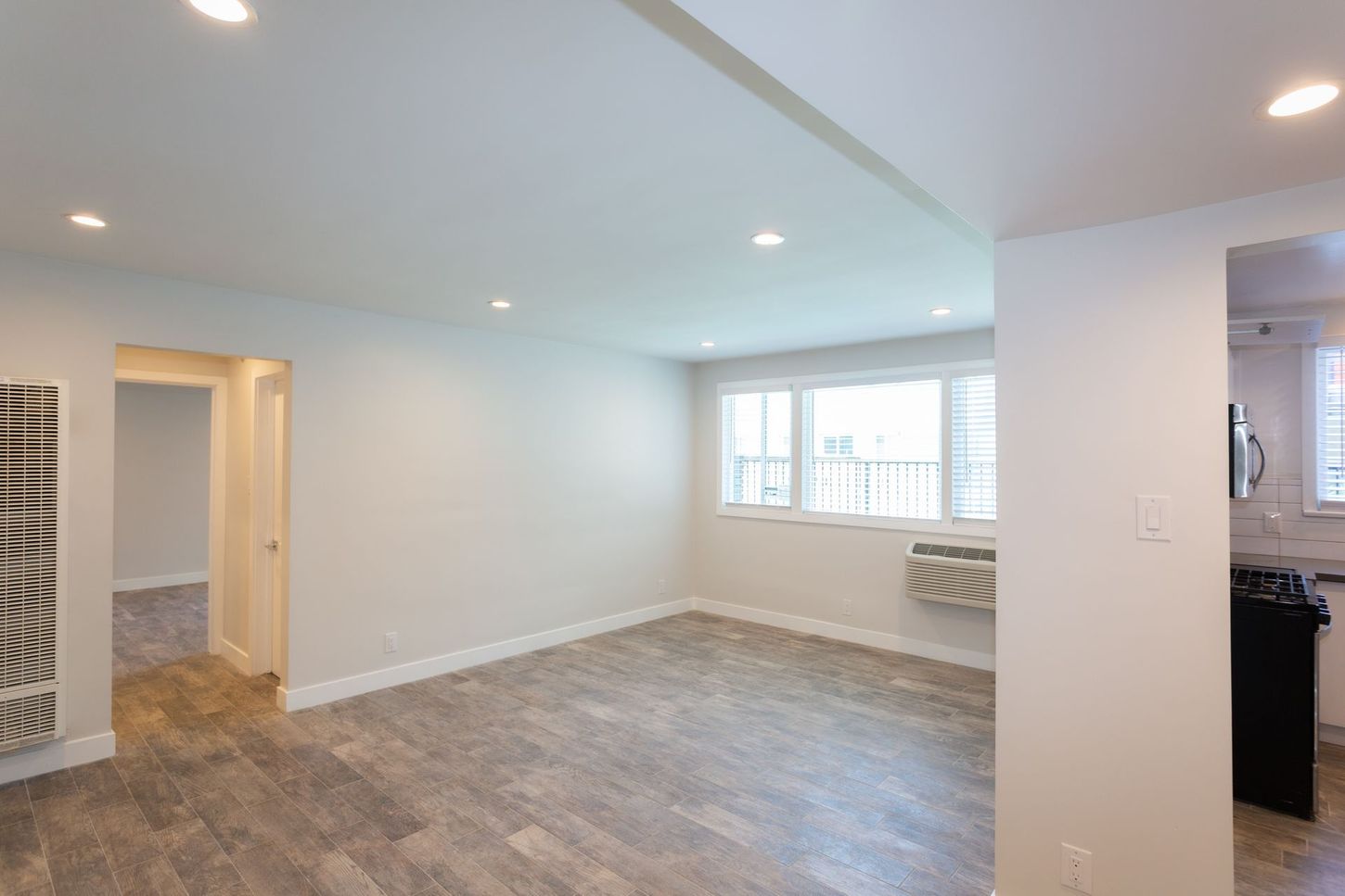 Empty living room with gray flooring, white walls, recessed lights, and a window by the kitchen pass-through.