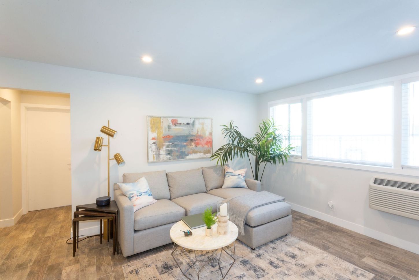 Bright living room with gray sectional sofa, round coffee table, plants, artwork, and large windows