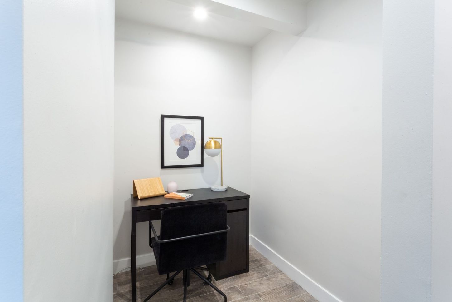 Small white home office with black desk, chair, lamp, and framed wall art.