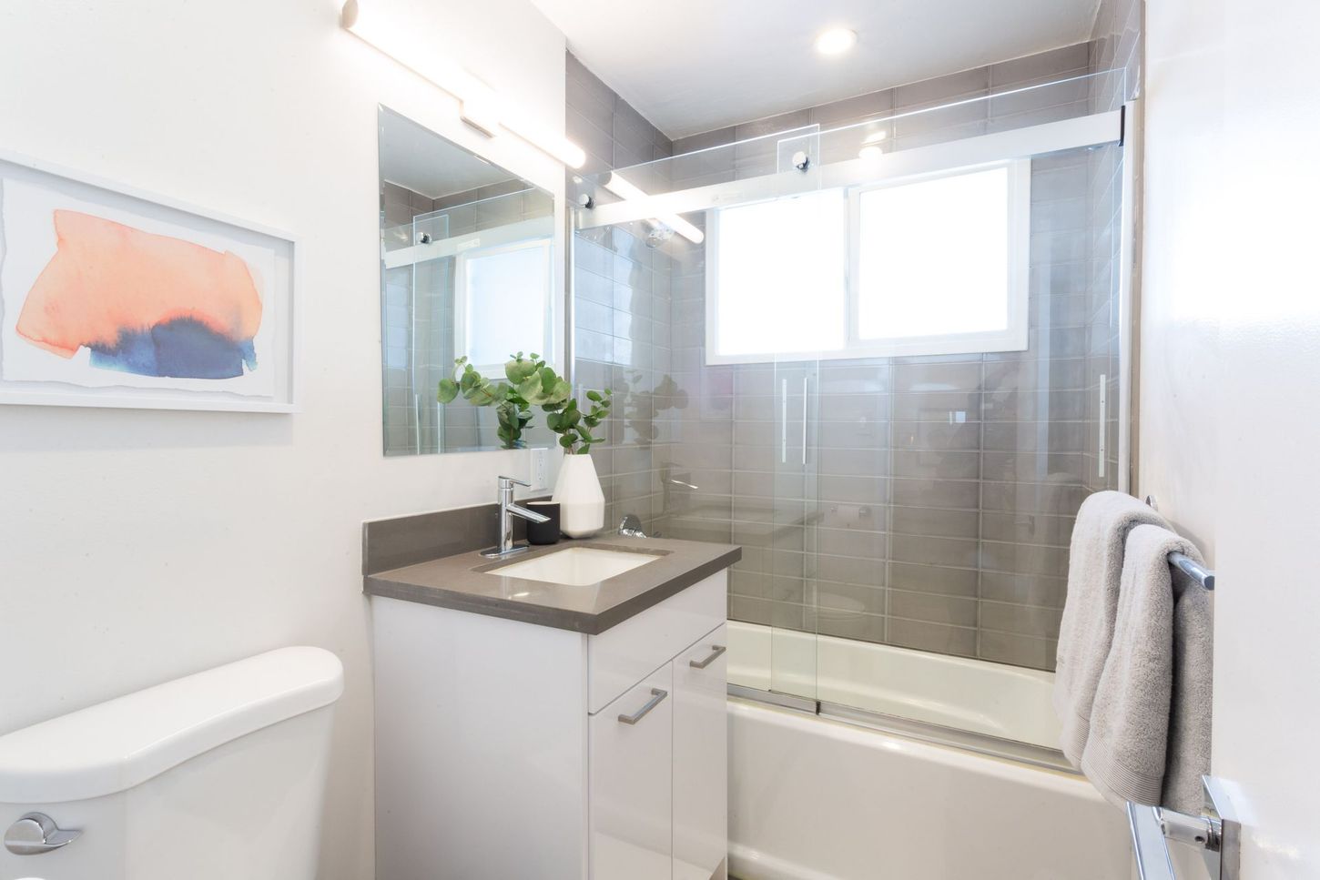 Bright white bathroom with bathtub, glass shower, vanity, toilet, and framed wall art