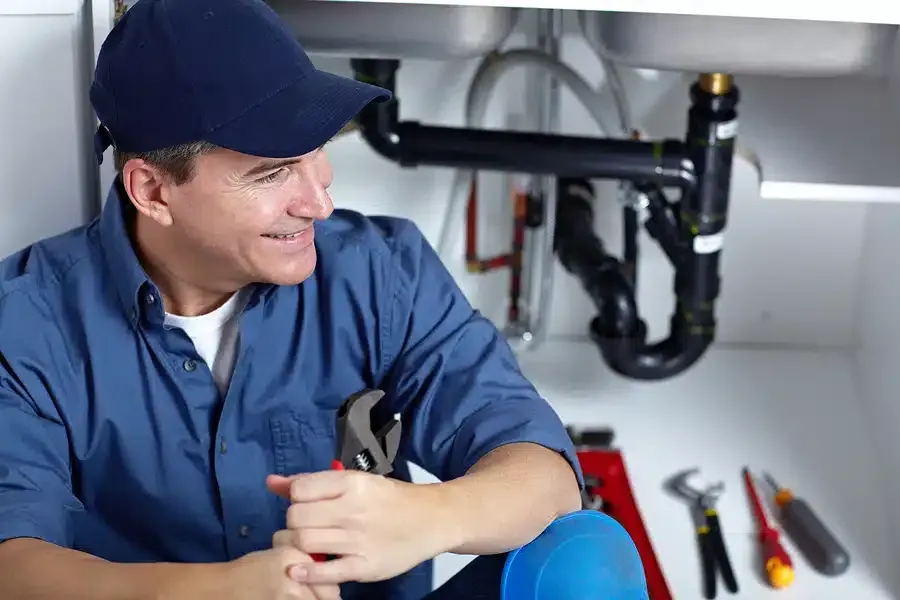 Plumber in blue shirt and hat smiling, holding pliers under a sink with tools.