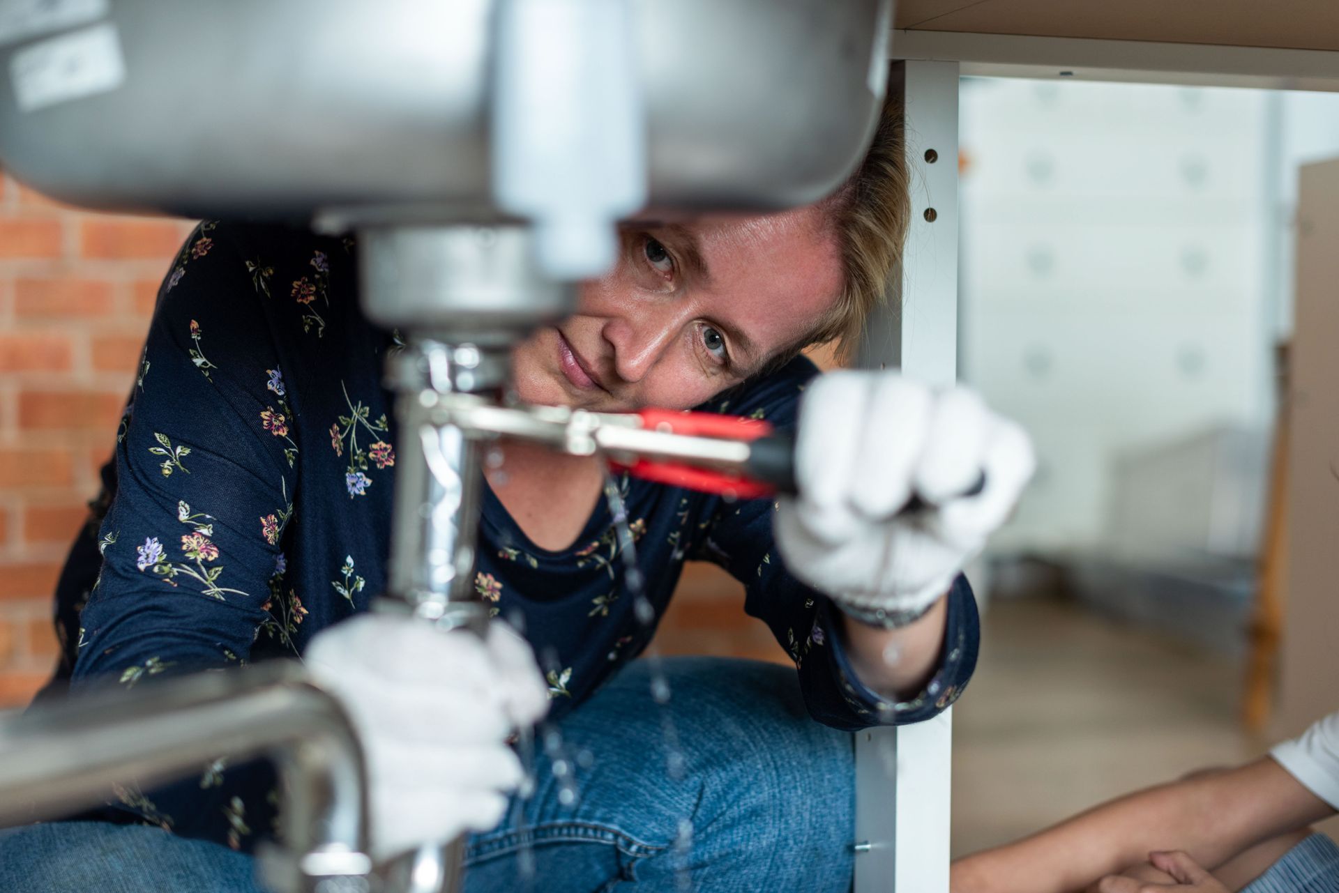 Person using a wrench to repair a pipe under a sink, wearing white gloves and a dark floral shirt.