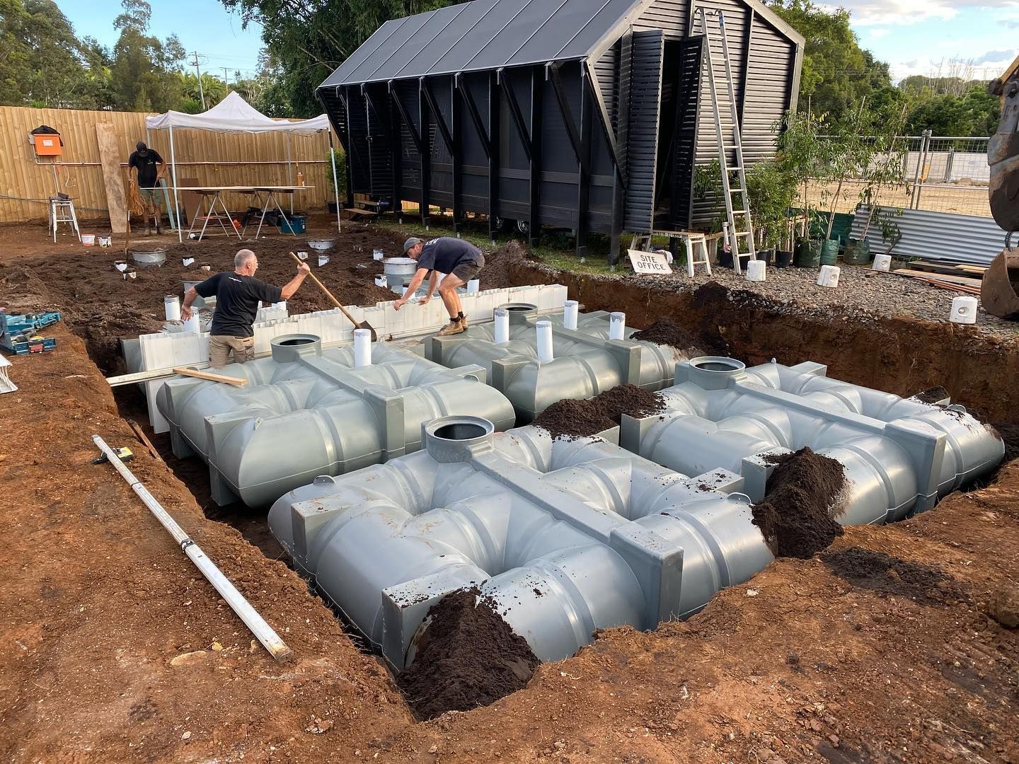 Workers installing gray plastic tanks in a dirt excavation, with a shed and fence in the background.