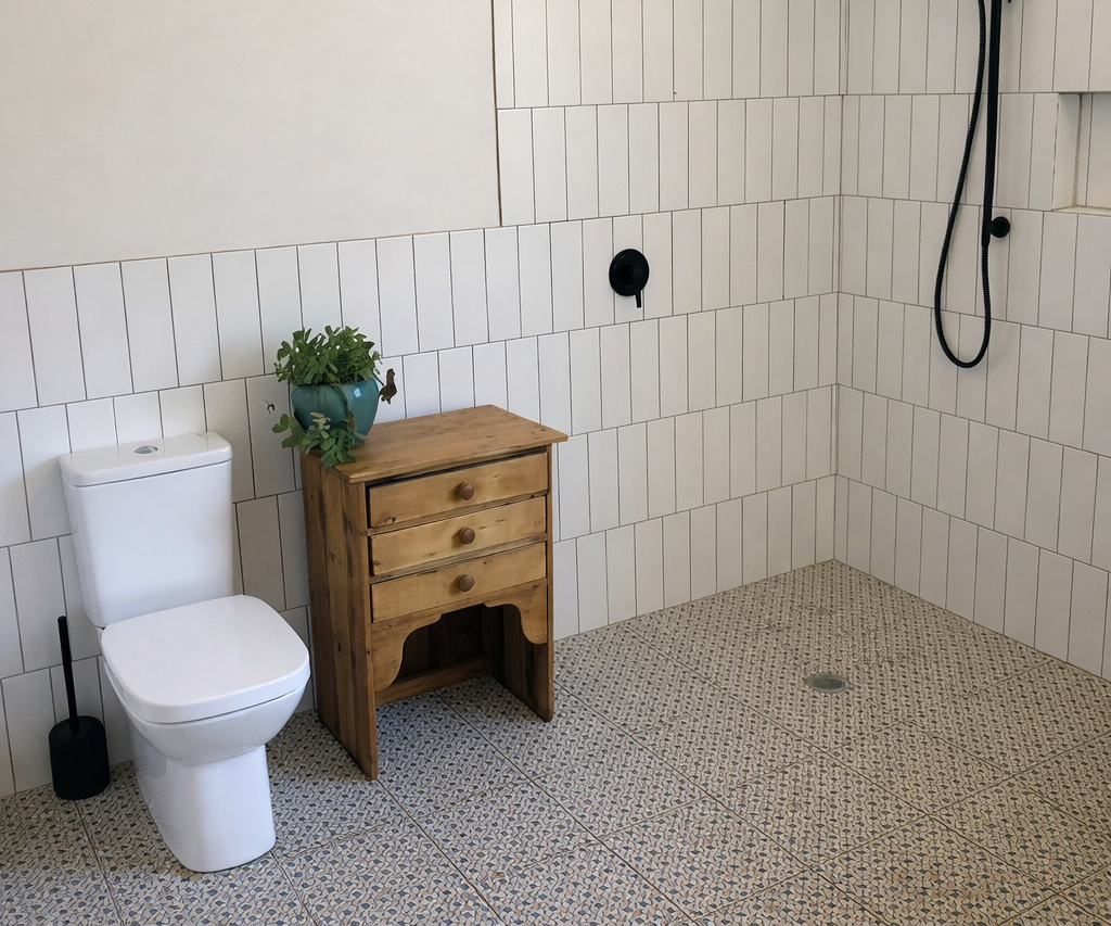 White-tiled bathroom with toilet, wooden cabinet, black shower head and fixture, and patterned floor.