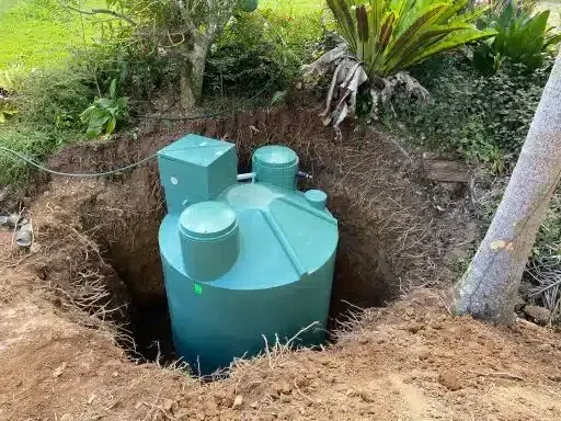 A green septic tank installed in a dug-out hole, surrounded by dirt, trees, and bushes.