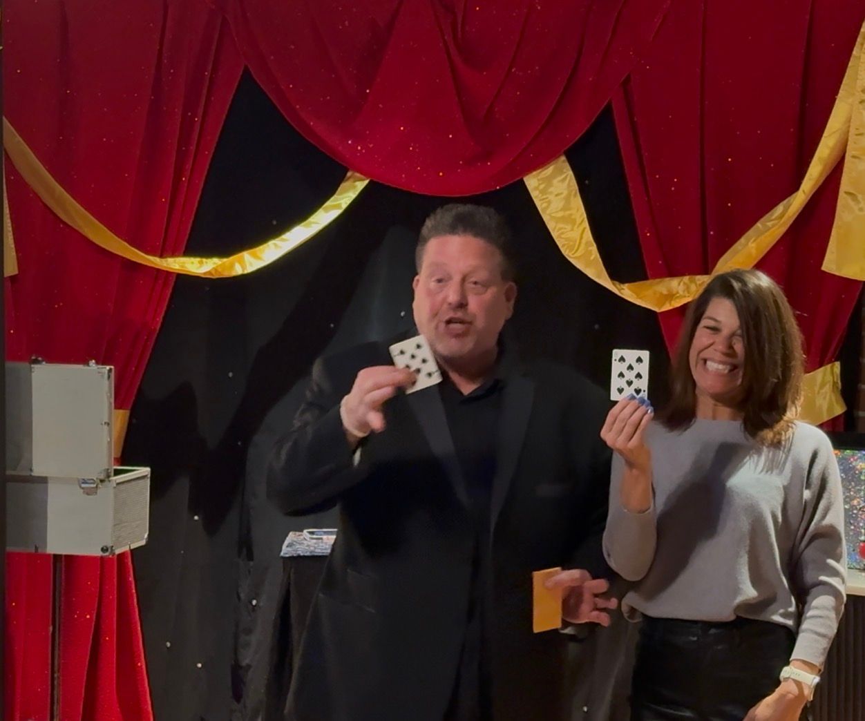 Two performers on stage holding playing cards before red and black drapes