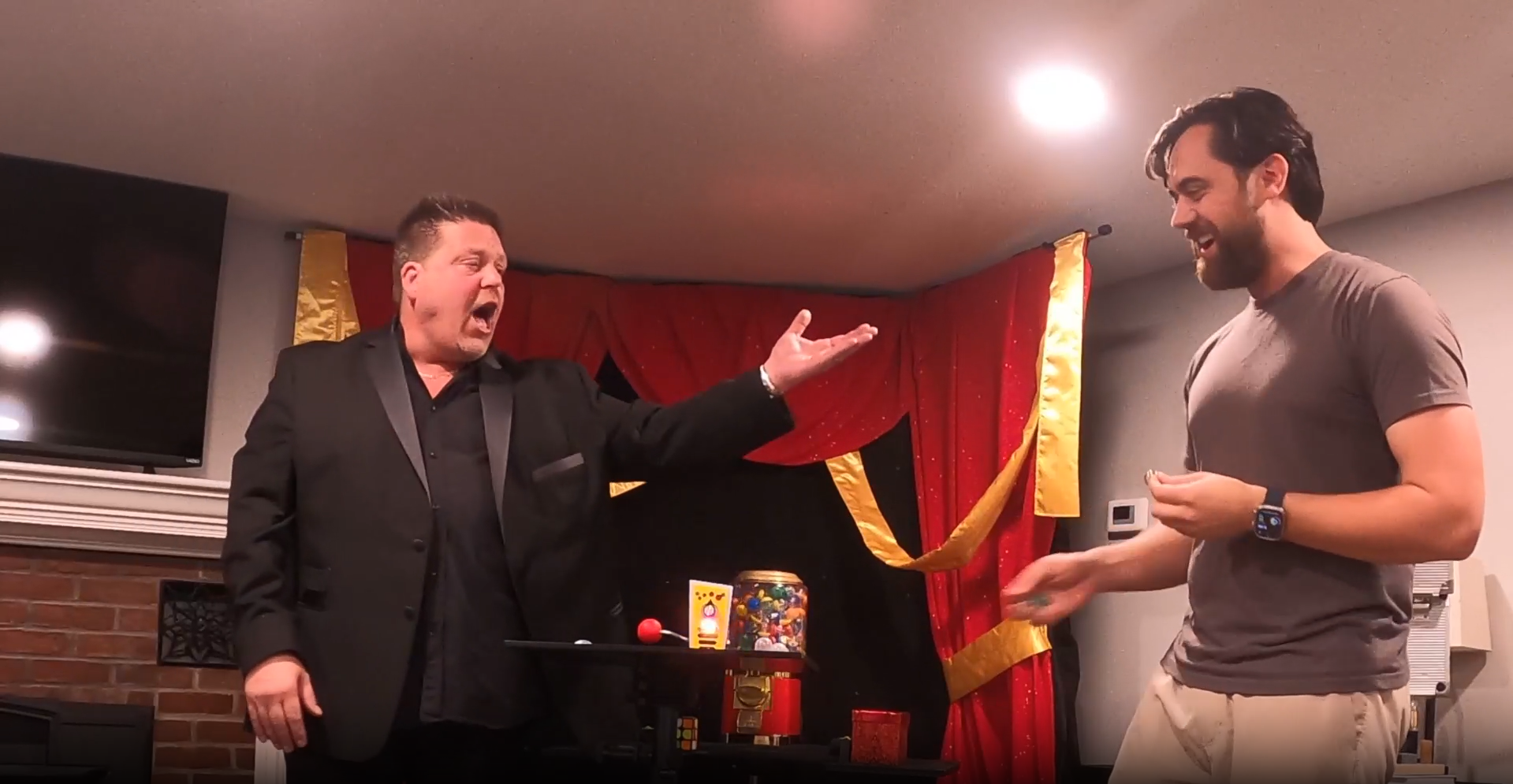 Two men talking in a dim room with red curtains and warm lighting, one gesturing broadly.
