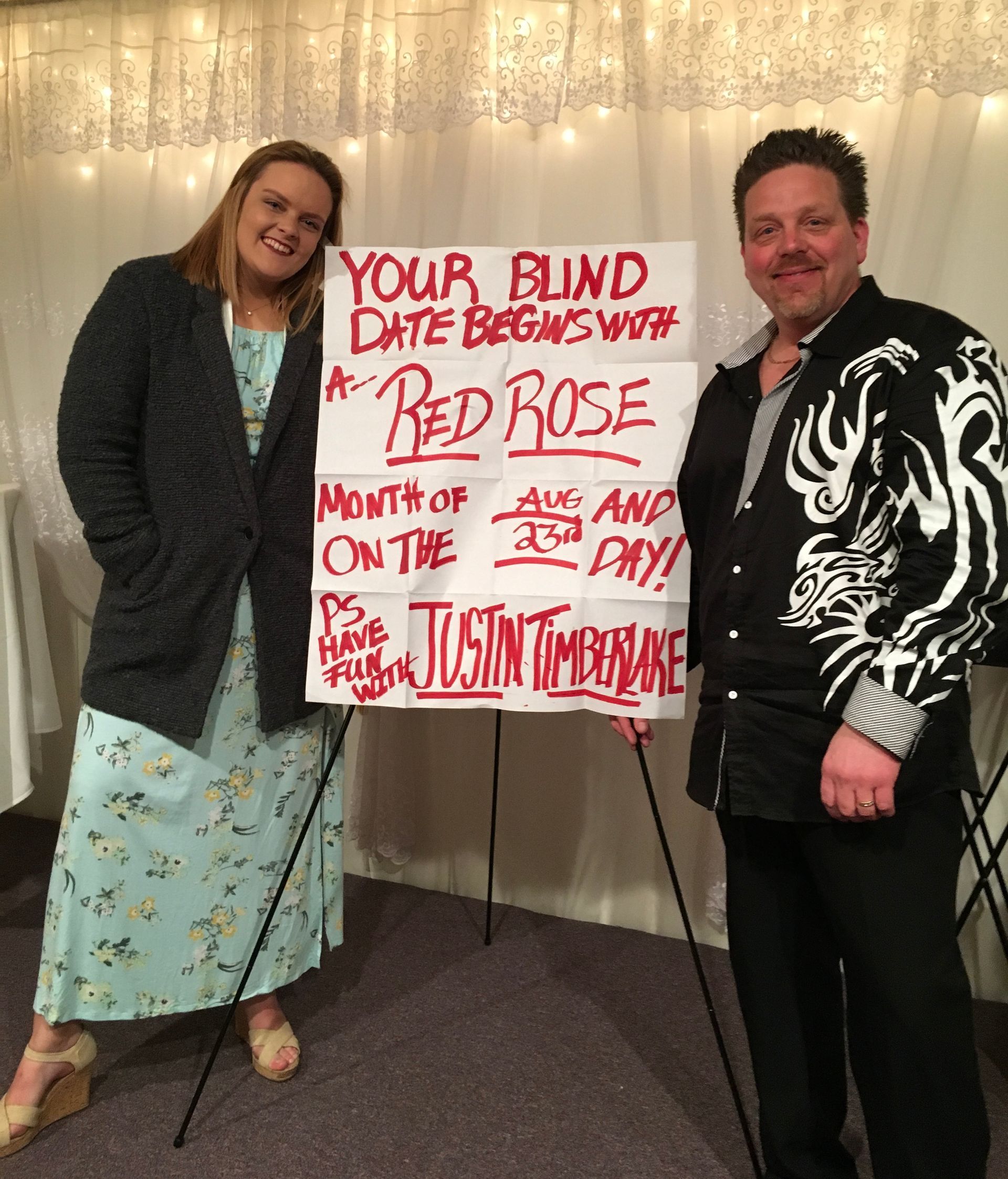 Two people posing beside a sign about “Bed Rose” at an indoor event.